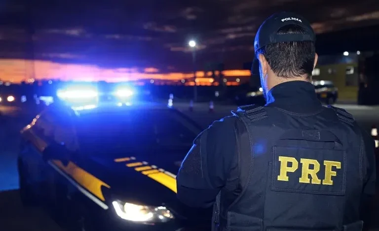 Agente da Polícia Rodoviária Federal (PRF), visto de costas e usando uniforme preto, aparece ao lado de uma viatura com luzes de emergência acionadas, durante a noite.