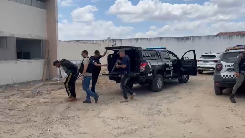 Policiais armados acompanham uma pessoa com a cabeça abaixada em um pátio, próximo a viaturas policiais estacionadas diante de um prédio