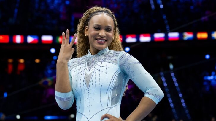 Rebeca Andrade sorri e ergue três dedos, vestindo collant azul claro no ginásio do Mundial