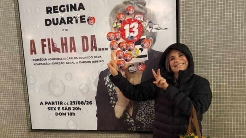 Regina Duarte faz o número 22 ao lado de cartaz de sua peça coberto por adesivos do PT