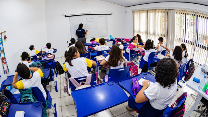 Estudantes uniformizados sentados em carteiras azuis acompanham professora diante do quadro