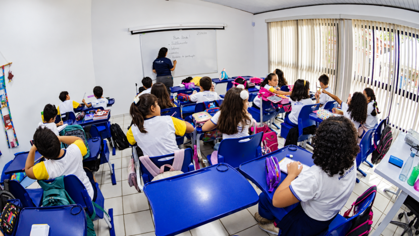 Estudantes uniformizados sentados em carteiras azuis acompanham professora diante do quadro