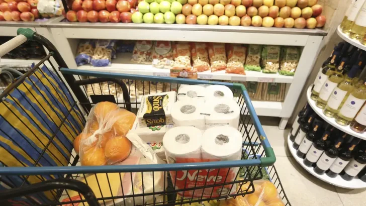 Carrinho de supermercado com frutas, embalagens de papel higiênico e outros produtos de consumo doméstico, em frente a uma banca de maçãs e prateleiras com alimentos e bebidas