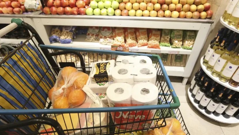 Carrinho de supermercado com frutas, embalagens de papel higiênico e outros produtos de consumo doméstico, em frente a uma banca de maçãs e prateleiras com alimentos e bebidas