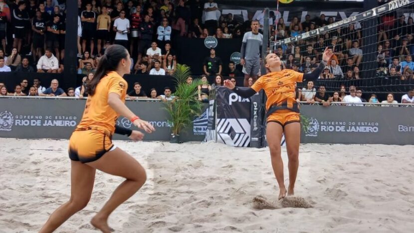 Duas atletas de uniforme laranja acompanham a bola junto à rede em quadra de areia, com público nas arquibancadas