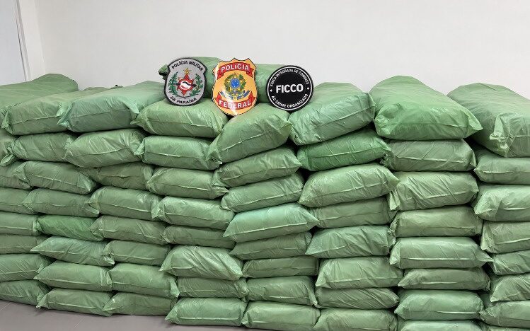 Dezenas de pacotes envolvidos em material verde aparecem empilhados diante de uma parede branca, com identificações da Polícia Federal, da Polícia Militar da Paraíba e da FICCO